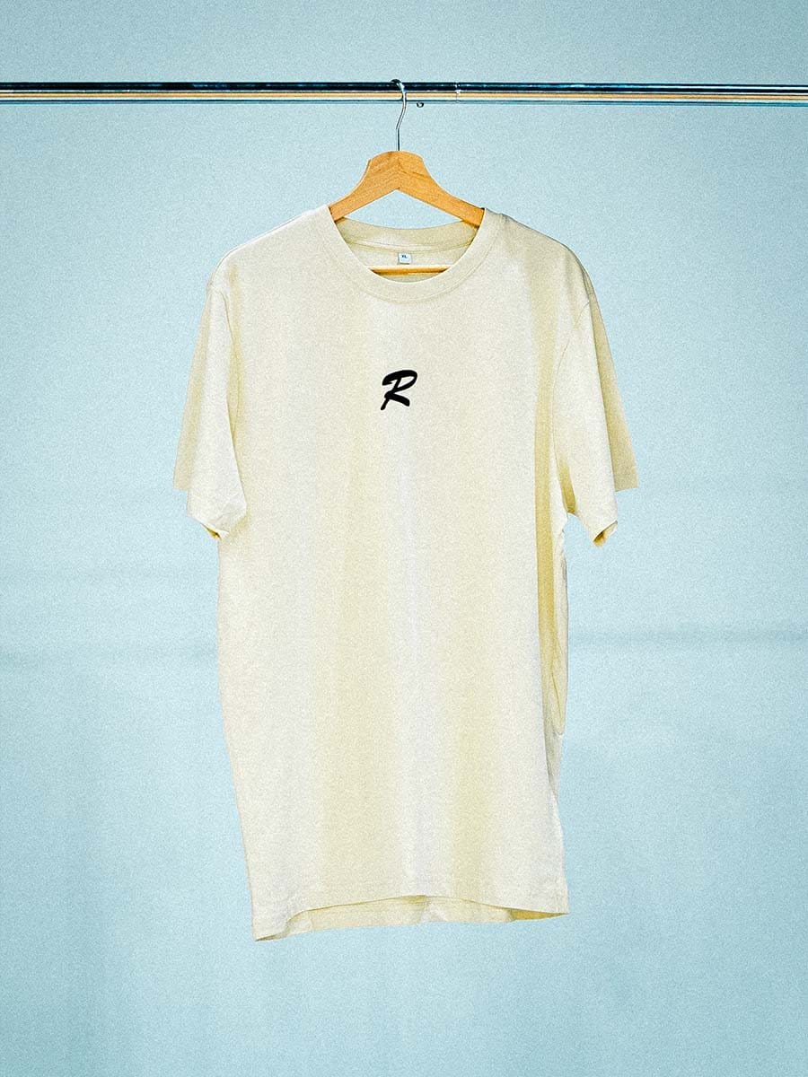 Helles T-Shirt mit schwarzem, kursivem Buchstaben "R" in der Mitte, hängt an einem Holzkleiderbügel an einer Stange vor einem hellblauen Hintergrund.