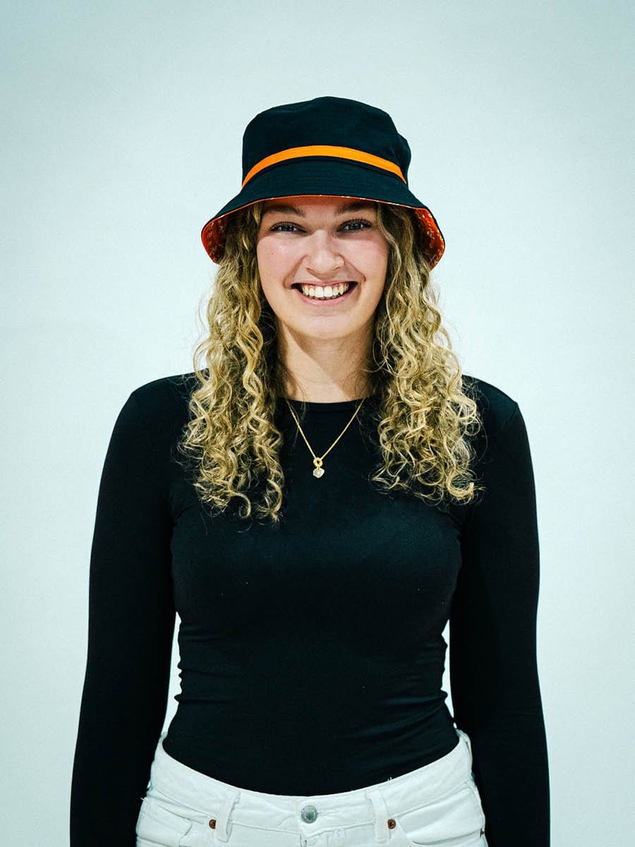 Junge Frau mit lockigem Haar, trägt schwarzen Hut mit orangefarbenem Band, schwarzes Oberteil und weiße Hose, lächelt vor hellem Hintergrund.