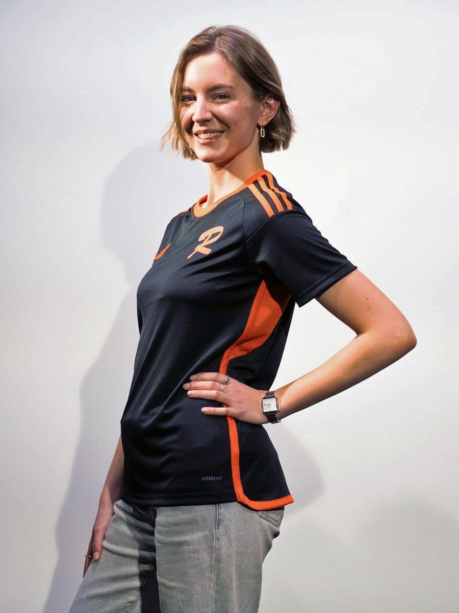 Junge Frau mit kurzen braunen Haaren, lächelnd, trägt ein schwarzes Sportshirt mit orangefarbenen Akzenten und graue Hosen, steht vor weißem Hintergrund.