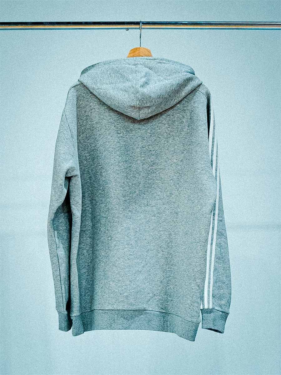 Grauer Kapuzenpullover mit weißen Streifen an den Ärmeln, hängt an einem Holzkleiderbügel an einer Stange vor einem hellblauen Hintergrund.