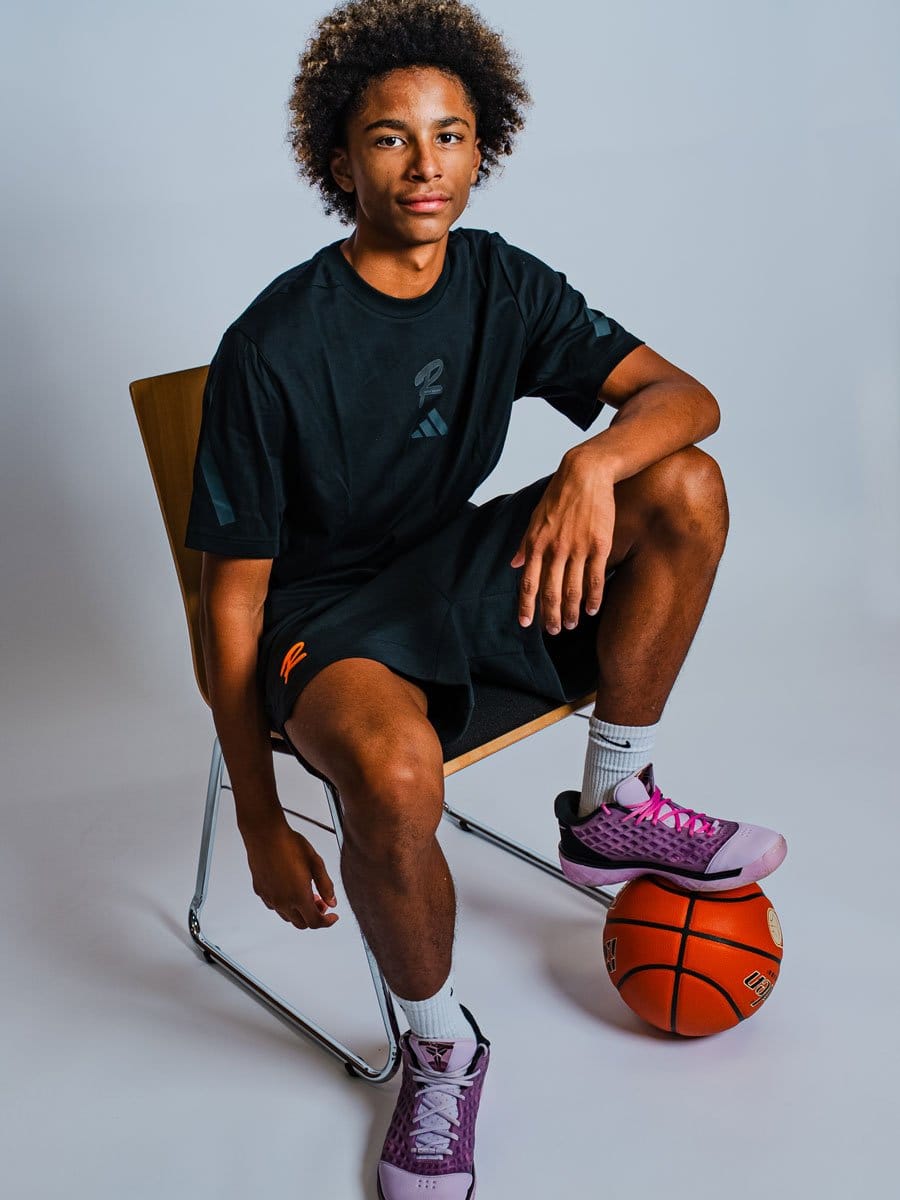 Junge Person sitzt auf einem Stuhl, hat ein Bein auf einem Basketball, trägt schwarze Sportkleidung und pink-lilafarbene Sportschuhe, vor einem grauen Hintergrund.