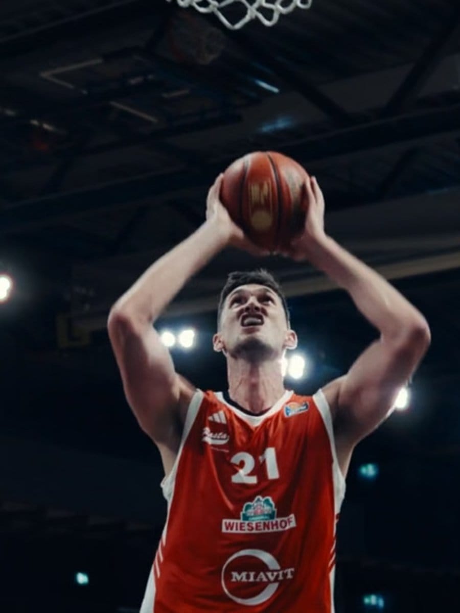 Basketballspieler in rotem Trikot mit der Nummer 21, springt zum Korb, hält den Ball hoch, im dunklen Indoor-Stadion mit Lichtern im Hintergrund.