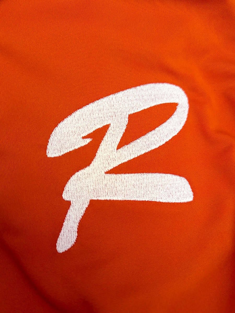Weißes, handgesticktes Logo in Form einer stilisierten Zahl 2 auf orangefarbenem Stoff.