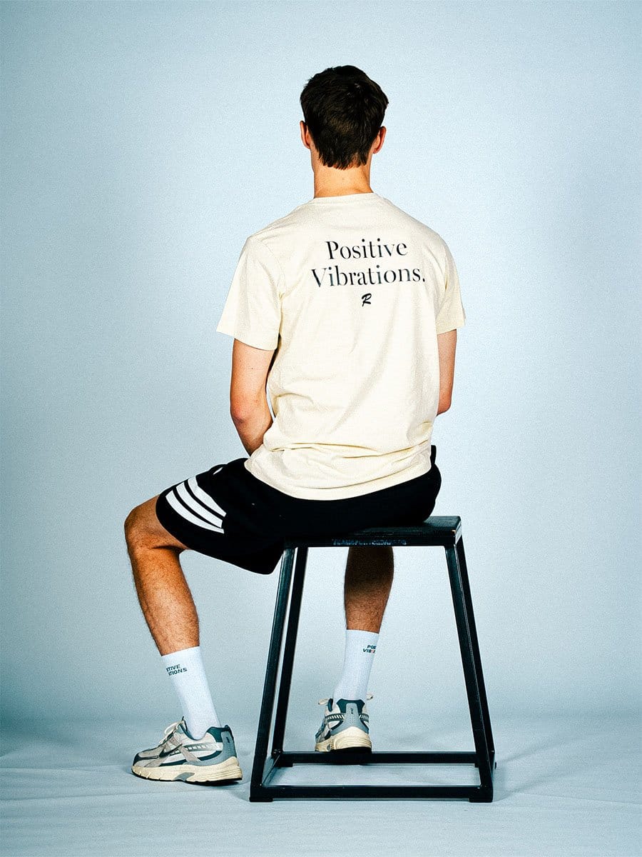 Junge Person sitzt auf einem schwarzen Hocker, trägt ein weißes T-Shirt mit "Positive Vibrations" auf dem Rücken, schwarze Shorts mit weißen Streifen, weiße Socken und Sneakers.