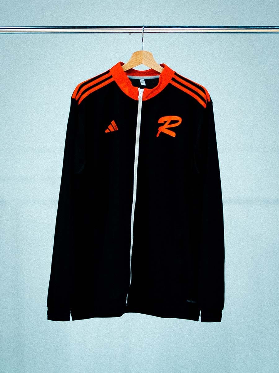 Schwarze Sportjacke mit roten Akzenten, Reißverschluss, Adidas-Logo und einem großen roten "R" auf der linken Brustseite, hängt an einem Holzkleiderbügel.