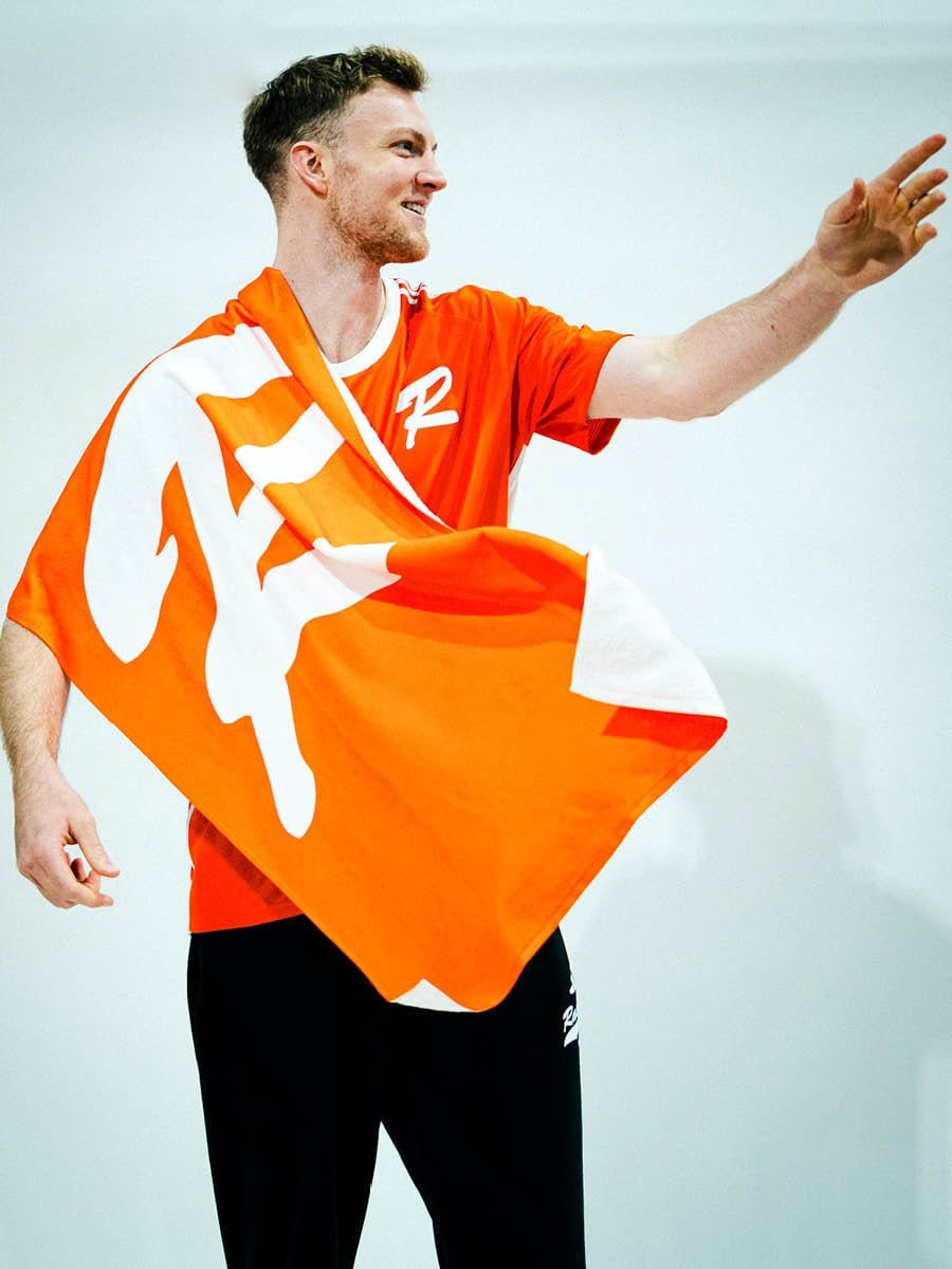 Junge Sportler hält eine orange und weiße Flagge, trägt ein rotes T-Shirt mit weißem Logo, lächelt und winkt, vor weißem Hintergrund.