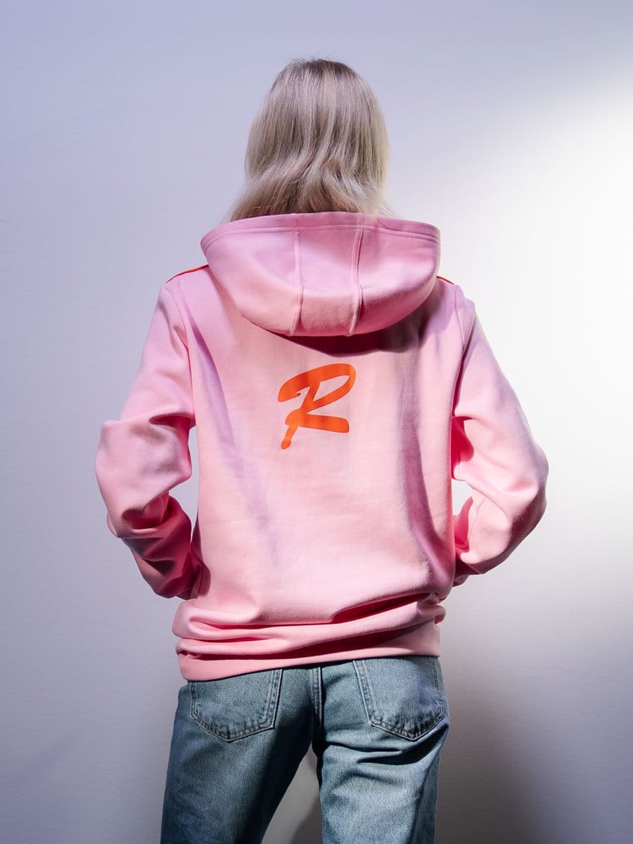 Person mit langem, blondem Haar trägt pinken Hoodie mit großem Logo auf dem Rücken, Jeans, steht vor weißer Wand, Rückenansicht.