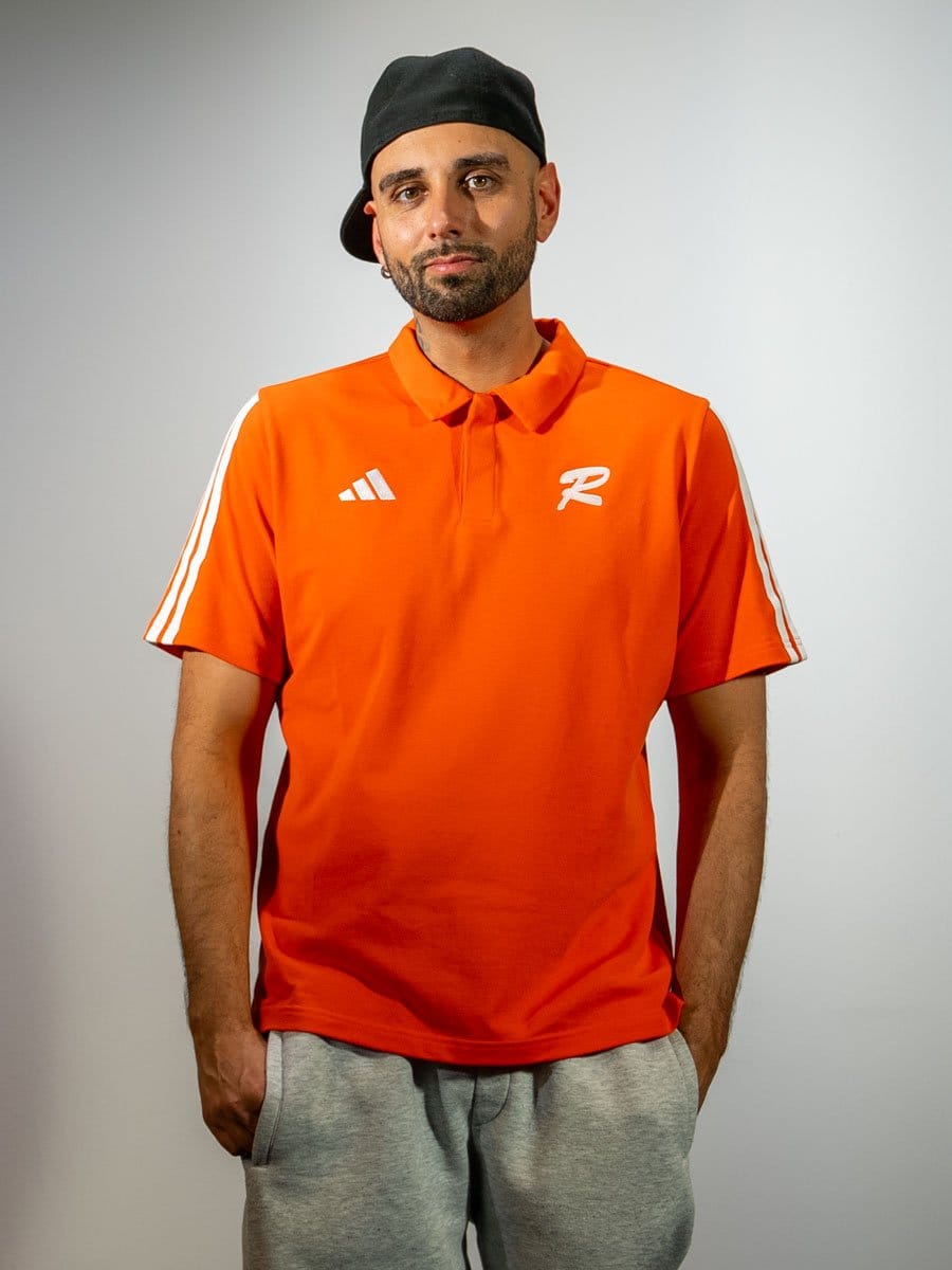 Mann trägt orange Polo-Shirt mit weißen Streifen an den Schultern, weißen Logos und graue Jogginghose, dazu schwarze Cap, blickt ernst in die Kamera.