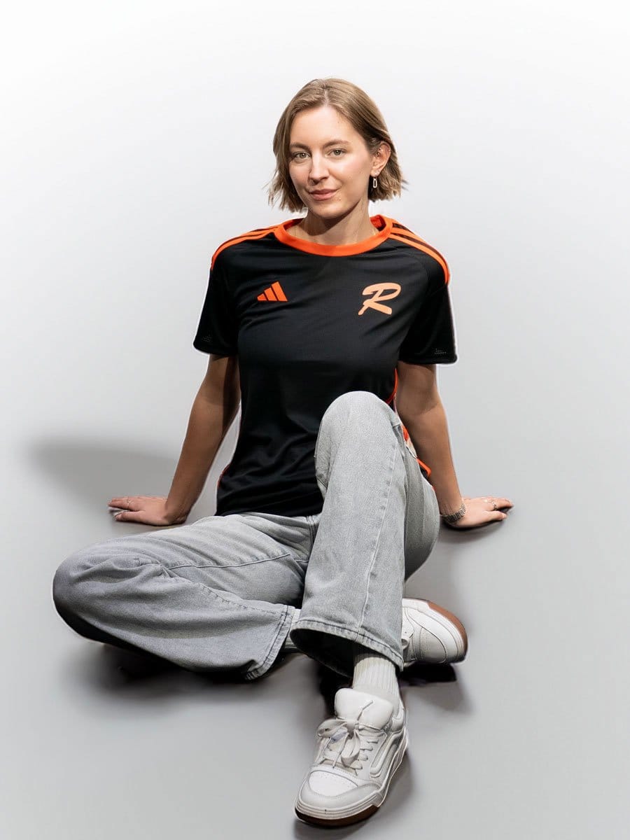 Junge Frau sitzt auf dem Boden, trägt ein schwarzes Sportshirt mit orangefarbenen Details, graue Jeans und weiße Sneakers, vor einem weißen Hintergrund.