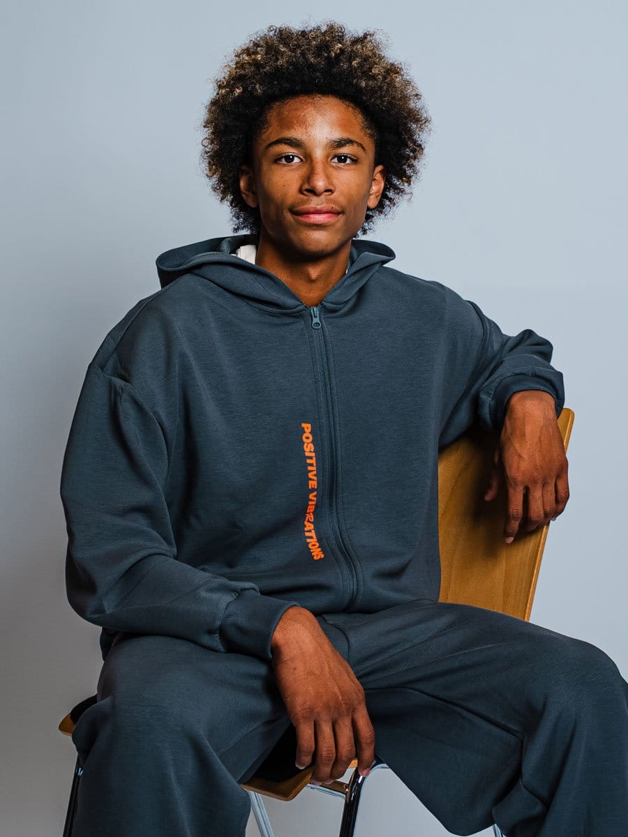 Junge Person mit dunkler Haut und lockigem Haar sitzt auf einem Stuhl, trägt eine dunkelblaue Kapuzenjacke mit orange Schriftzug "POSITIVE VIBRATIONS" und blickt in die Kamera.