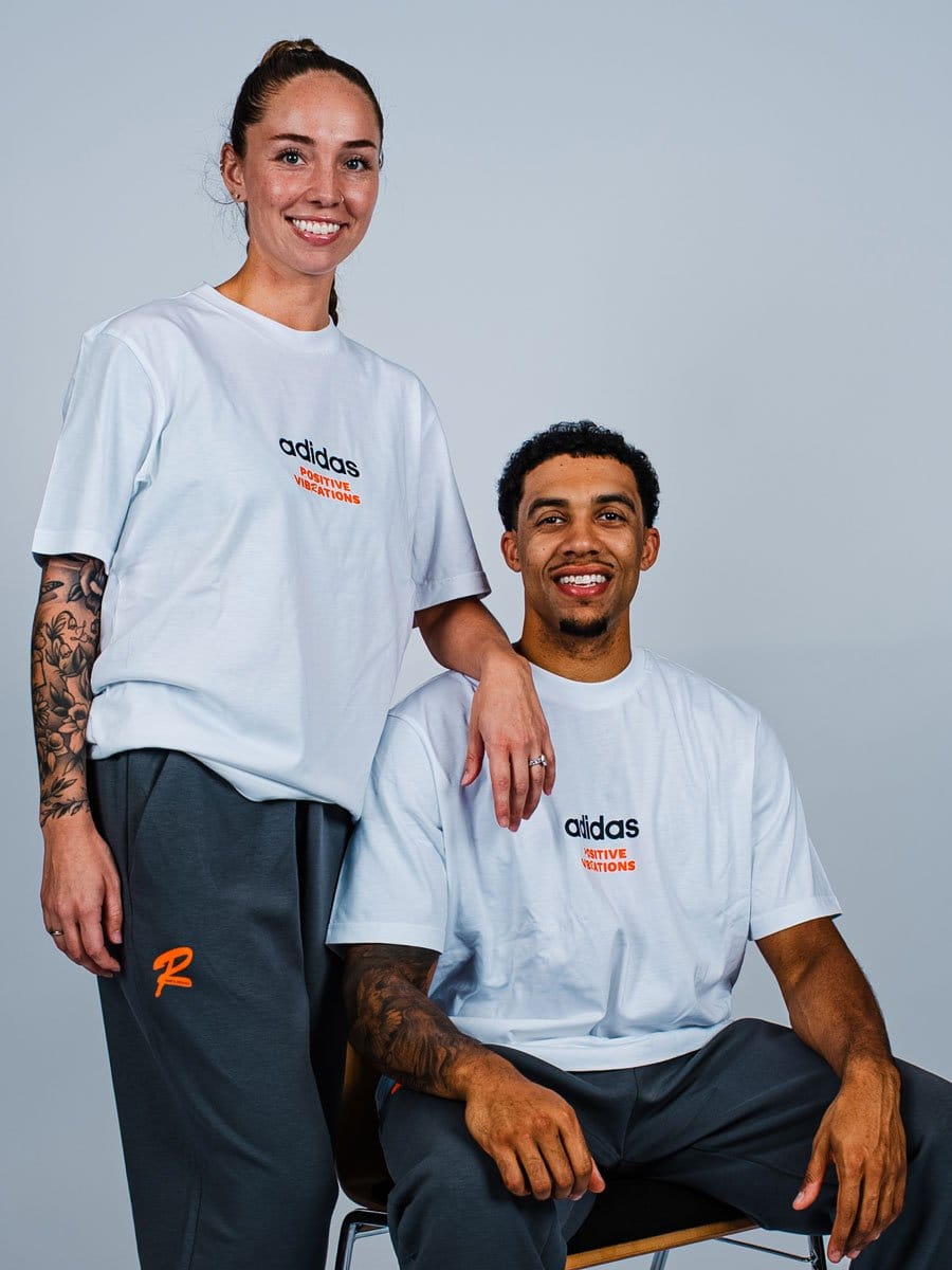 Ein lächelndes Paar in weißen Adidas-T-Shirts mit "Positive Vibrations" Logo, die Frau steht, der Mann sitzt auf einem Stuhl, beide vor hellem Hintergrund.