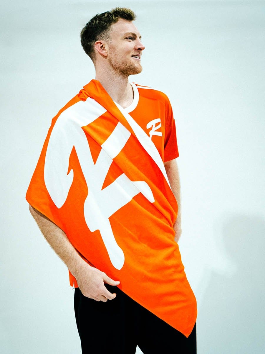 Junge Mann in orange und weißem Sportshirt, hält ein großes orange und weißes Tuch, steht vor hellem Hintergrund, lächelt leicht, Blick nach rechts.