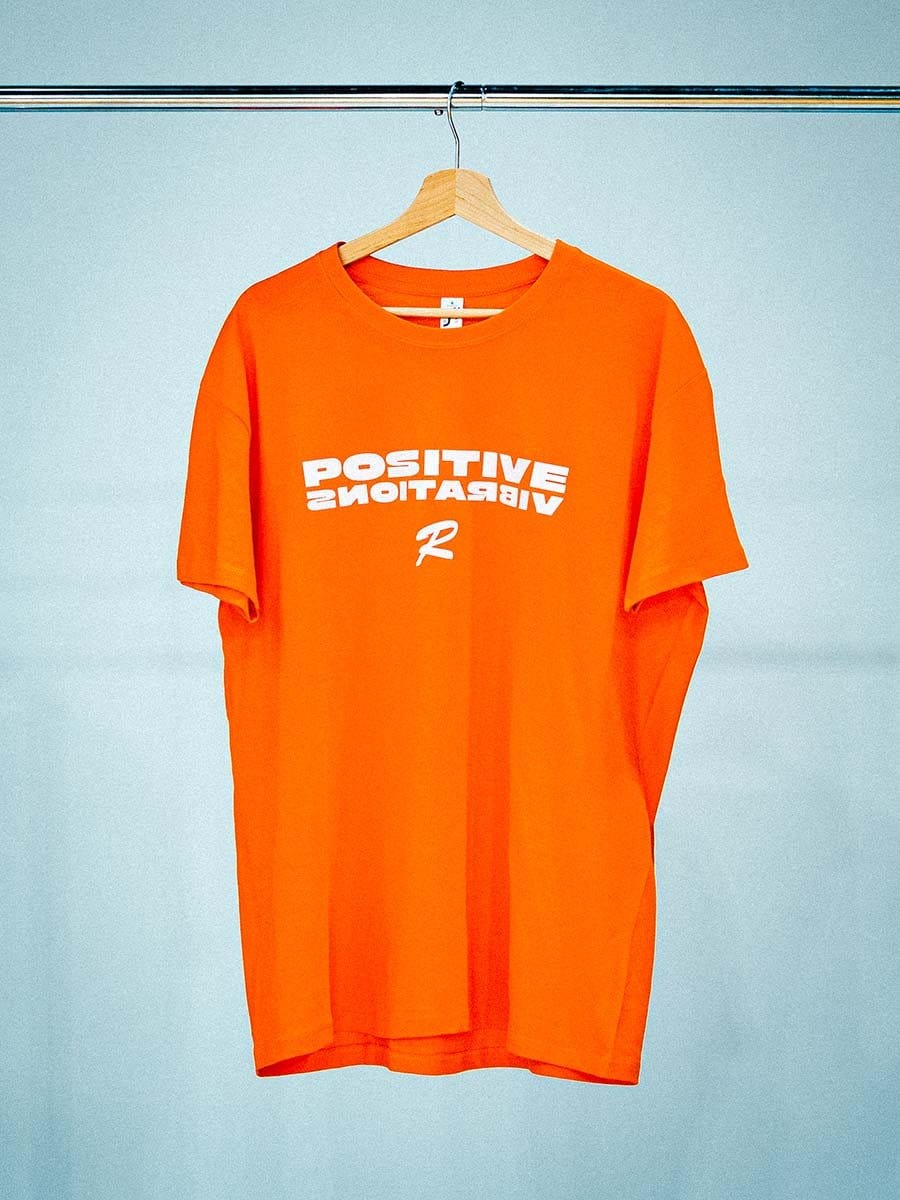 Oranges T-Shirt mit weißen "Positive Vibrations" Schriftzug und einem weißen "R" Logo, hängt an einem Holzkleiderbügel an einer Stange.