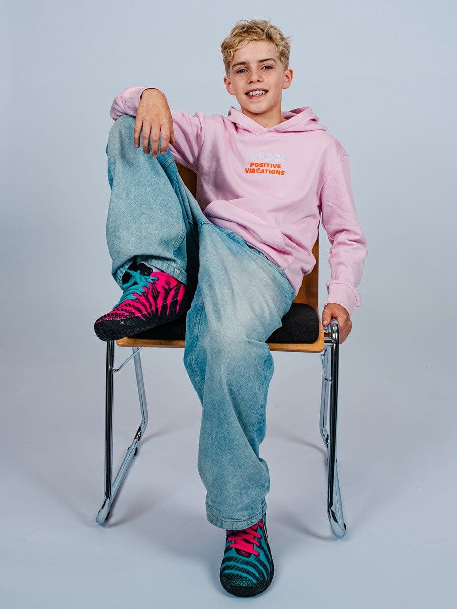 Junge sitzt auf einem Stuhl, trägt eine pinke Hoodie mit "Positive Vibrations" Schrift, blaue Jeans und bunte Sportschuhe, lächelt freundlich.