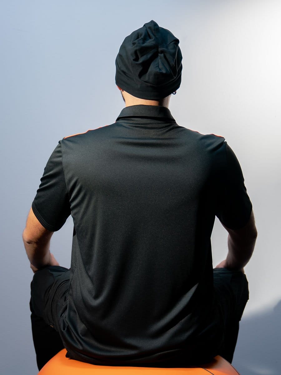 Person sitzt mit Rücken zur Kamera, trägt schwarze Kleidung und einen schwarzen Hut, sitzt auf einem orangefarbenen Sitz, vor einem hellen Hintergrund.