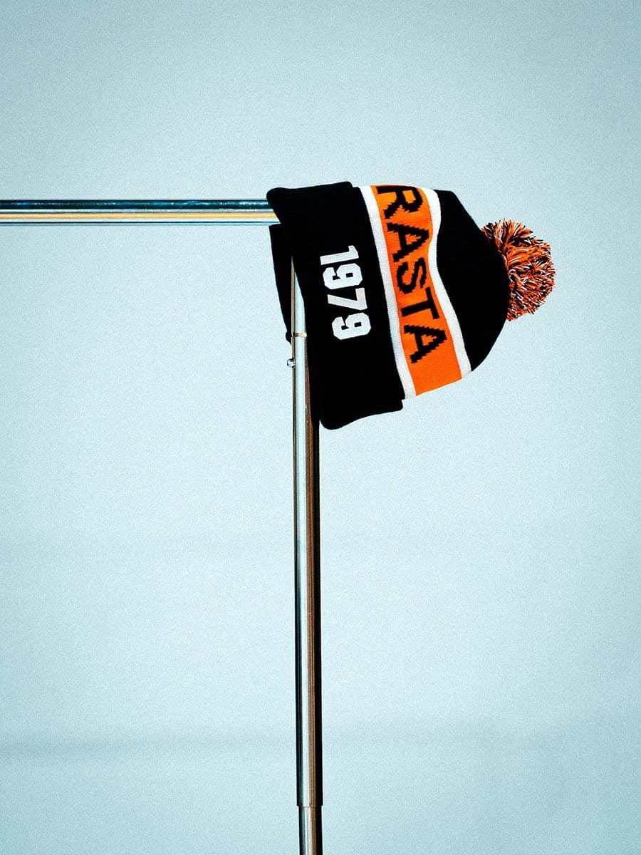 Schwarze Mütze mit orange-weißer Banderole und Pompon, hängt an einem Metallständer gegen einen hellblauen Hintergrund.