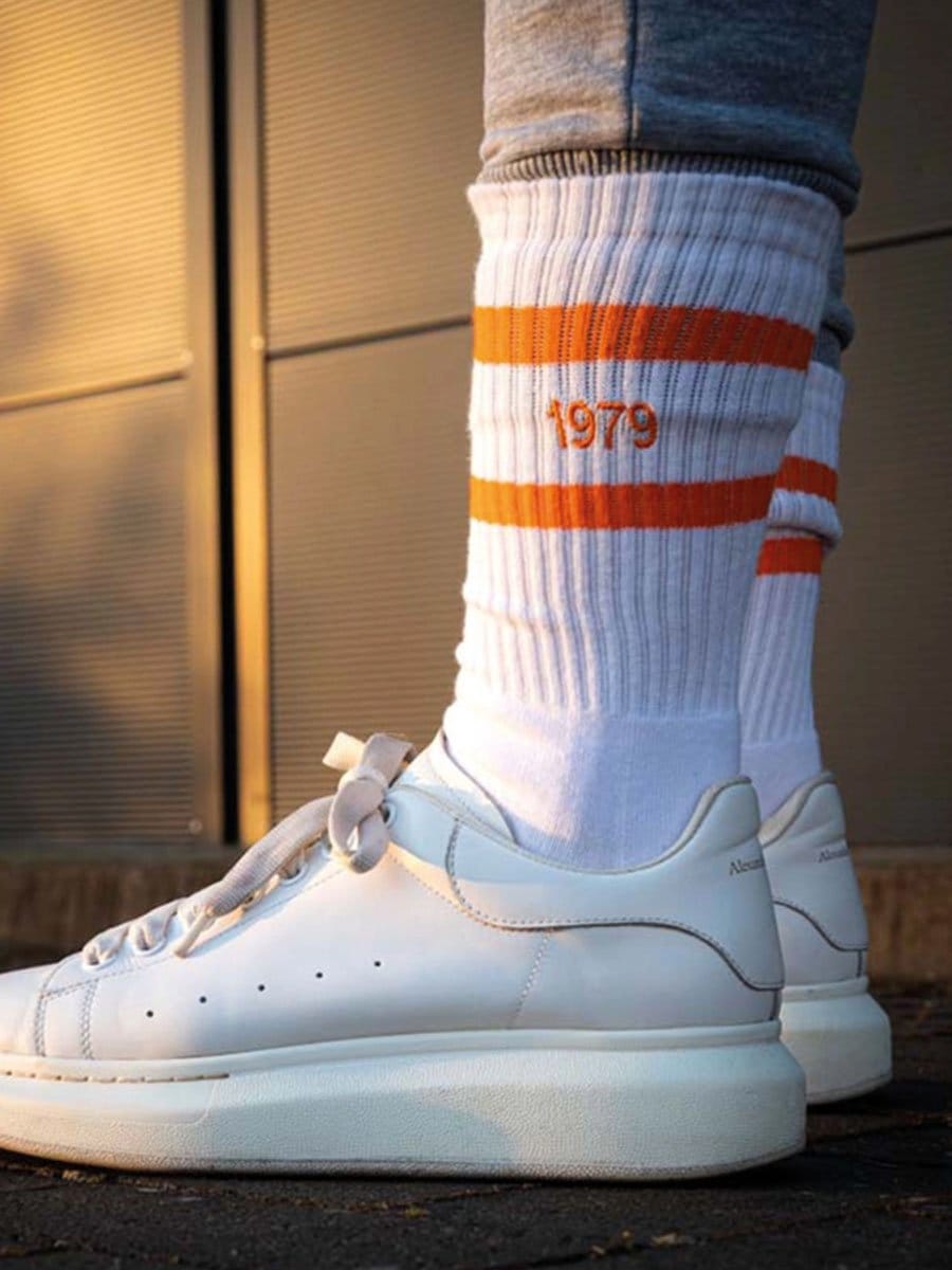 Weißer Sneaker mit dicker Sohle, getragen mit weißen Socken mit orangefarbenen Streifen und der Jahreszahl 1979, vor einem modernen Gebäude.