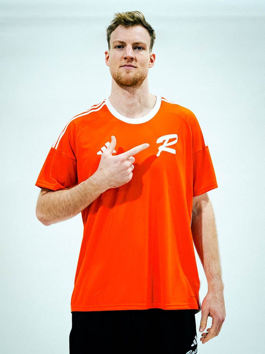 Junger Mann in rotem Sportshirt mit weißen Streifen und Logo, zeigt auf die Brust, steht vor weißem Hintergrund, blickt ernst in die Kamera.