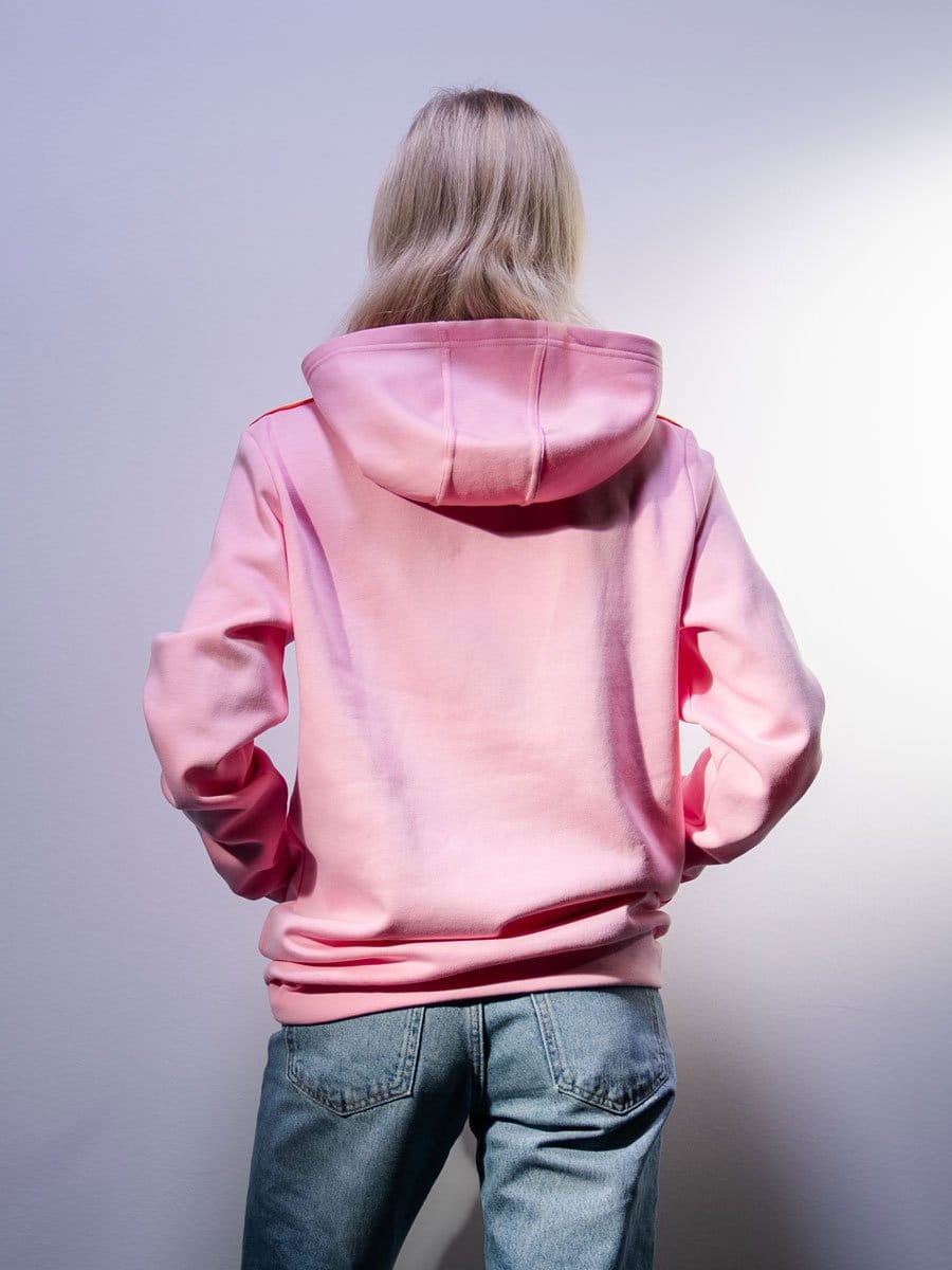 Junge Frau mit langem, blondem Haar trägt eine pinke Kapuzenjacke und blaue Jeans, steht mit dem Rücken zur Kamera vor einer hellen Wand.