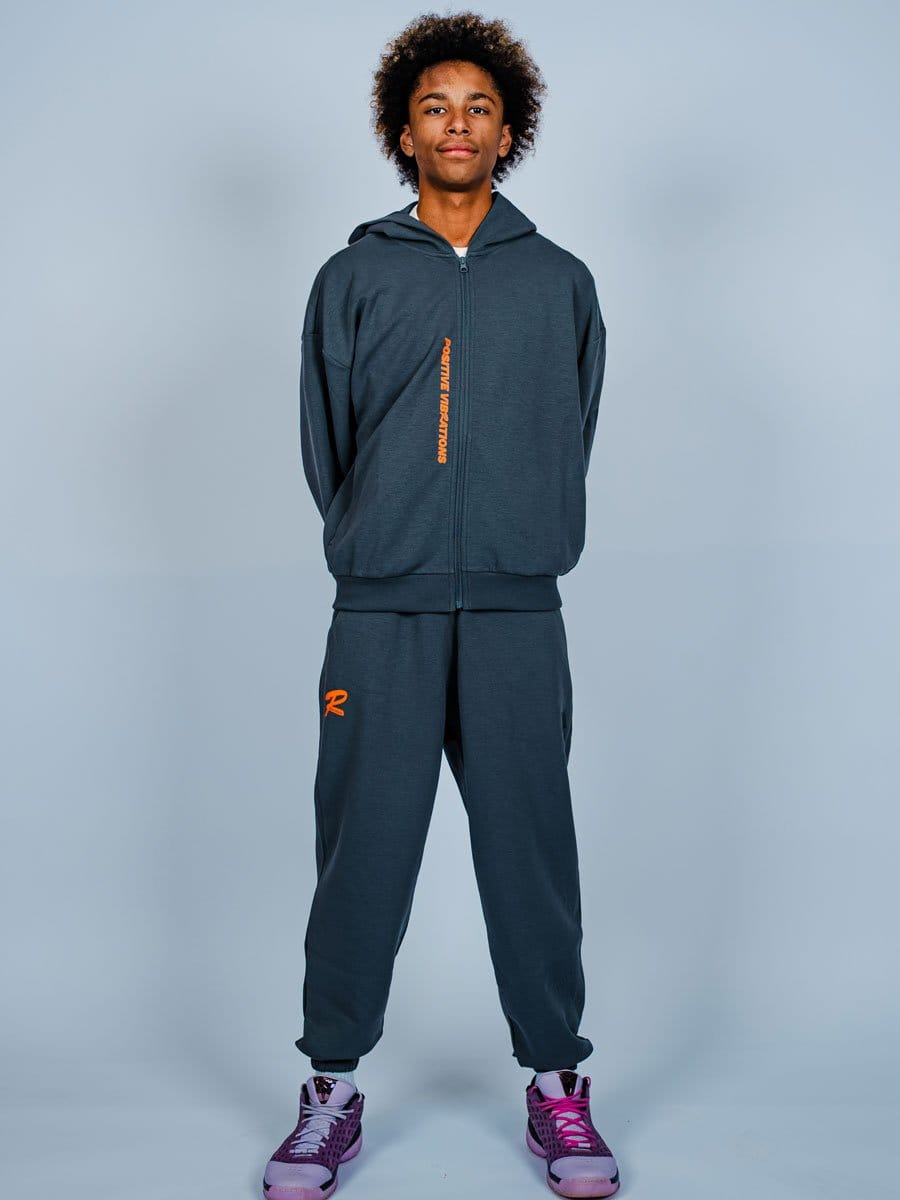 Junge Person in dunklem Sportoutfit mit Hoodie und Jogginghose, trägt pink-lila Sportschuhe, steht vor hellem Hintergrund, wirkt selbstbewusst.