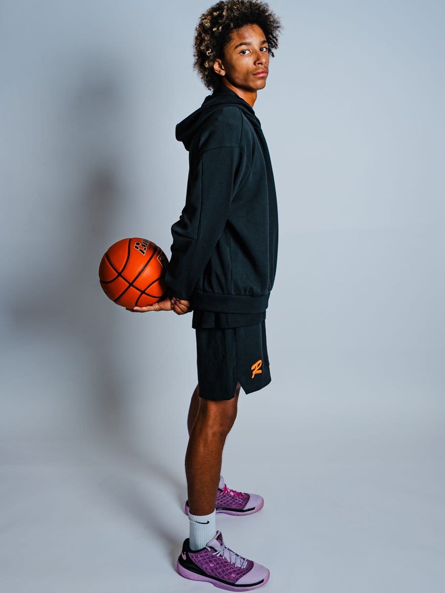 Junge Basketballspieler in schwarzer Kapuzenjacke, Shorts und pinken Sportschuhen, hält einen Basketball hinter dem Rücken, blickt ernst in die Kamera.