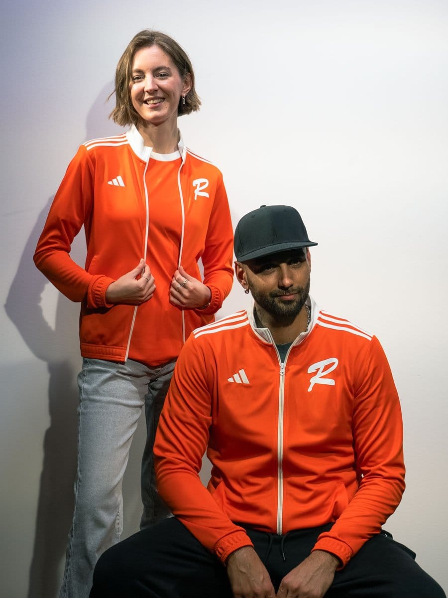 Ein Mann und eine Frau tragen beide orange Sportjacken mit weißen Streifen und Logos, die vor einem weißen Hintergrund stehen.