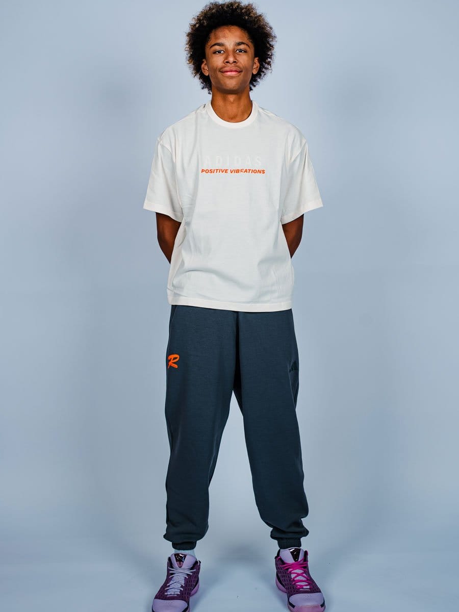Junge Person in weißem T-Shirt mit orange Schrift, dunklen Jogginghosen und lila Turnschuhen, steht vor hellem Hintergrund, lächelt leicht.