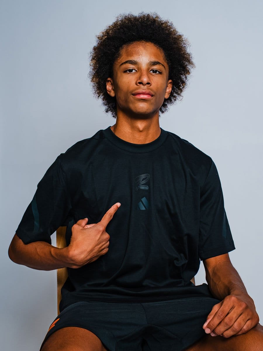 Junger Mann mit Afro-Haar, sitzend auf einem Stuhl, trägt schwarze Sportkleidung, zeigt auf ein Logo auf seinem T-Shirt, vor hellem grauem Hintergrund.
