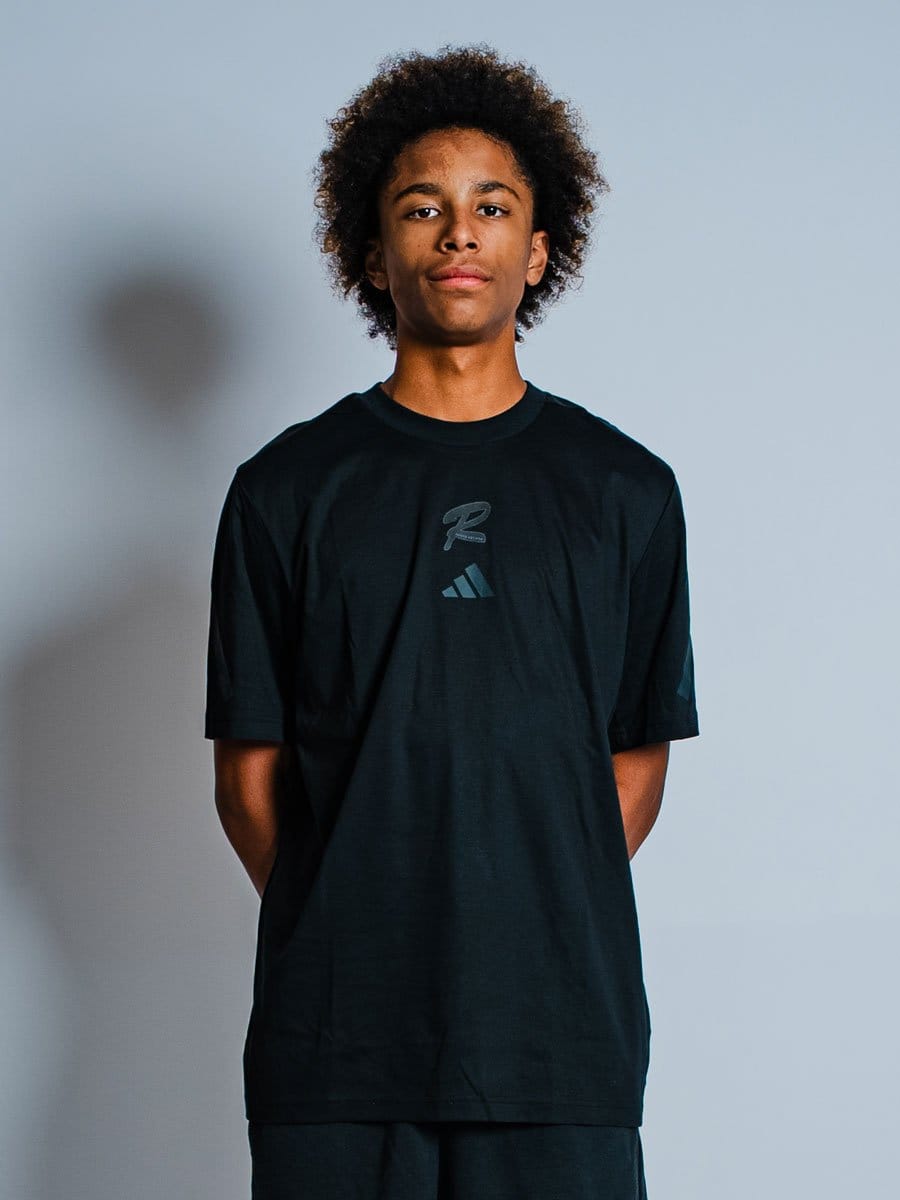 Junge Person mit Afro-Haar trägt schwarzen Adidas-Sport-T-Shirt, steht vor hellem Hintergrund, Blick direkt in die Kamera, Hände hinter dem Rücken.