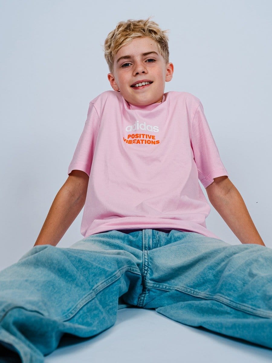 Junge mit blonden Haaren sitzt im Sitzen, trägt ein pinkes T-Shirt mit weißen und orangefarbenen Schriftzug, blaue Jeans, vor hellem Hintergrund.