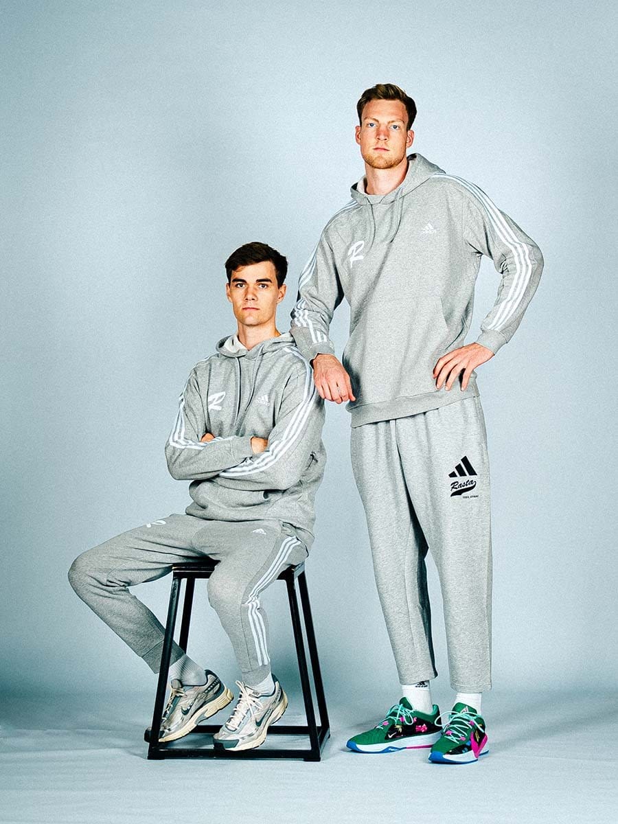 Zwei junge Männer in grauen Adidas-Sportoutfits mit weißen Streifen, sitzend und stehend vor einem hellblauen Hintergrund, tragen sportliche Sneakers.