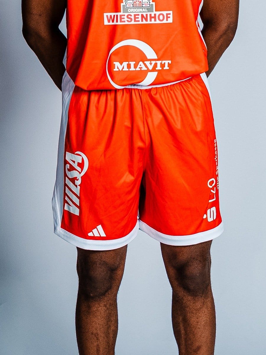 Blick auf die unteren Körperhälfte eines Mannes in roten Sportshorts mit weißen Seitenstreifen und Logos, gegen einen grauen Hintergrund.