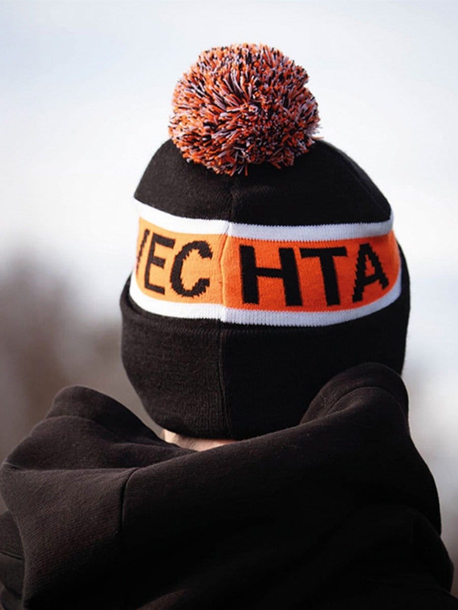 Mütze mit buntem Pompon, schwarzer Grundfarbe, orange-weißer Streifen mit schwarzen Buchstaben, trägt eine Person mit schwarzem Hoodie.
