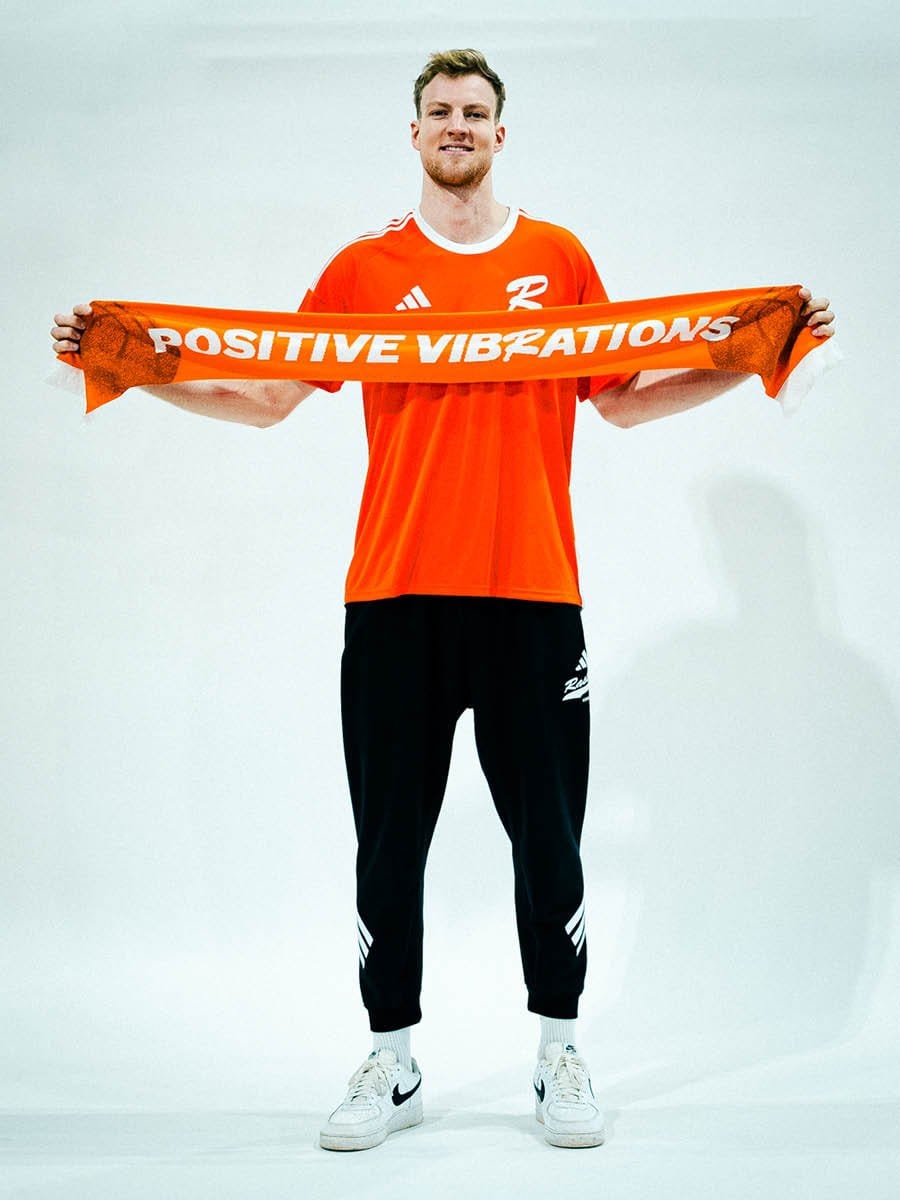 Junger Mann in orange Sportshirt und schwarzen Jogginghosen hält einen orangefarbenen Schal mit weißen Buchstaben, auf dem "POSITIVE VIBRATIONS" steht, vor einer weißen Wand.