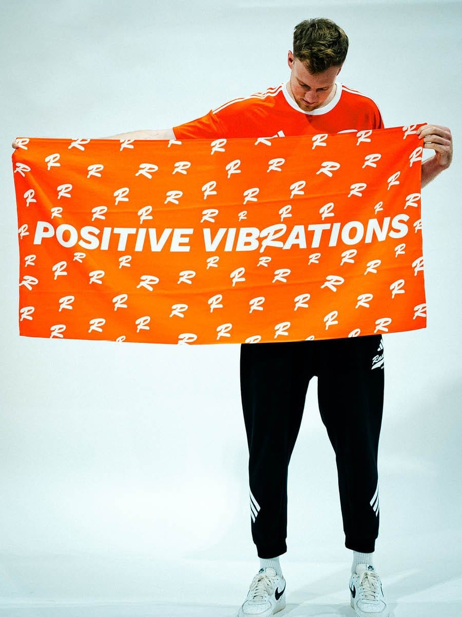 Junge Person hält ein orangefarbenes Tuch mit weißen Schriftzug "POSITIVE VIBRATIONS" und kleinen weißen R-Logos, trägt schwarze Jogginghose und weiße Sneakers.