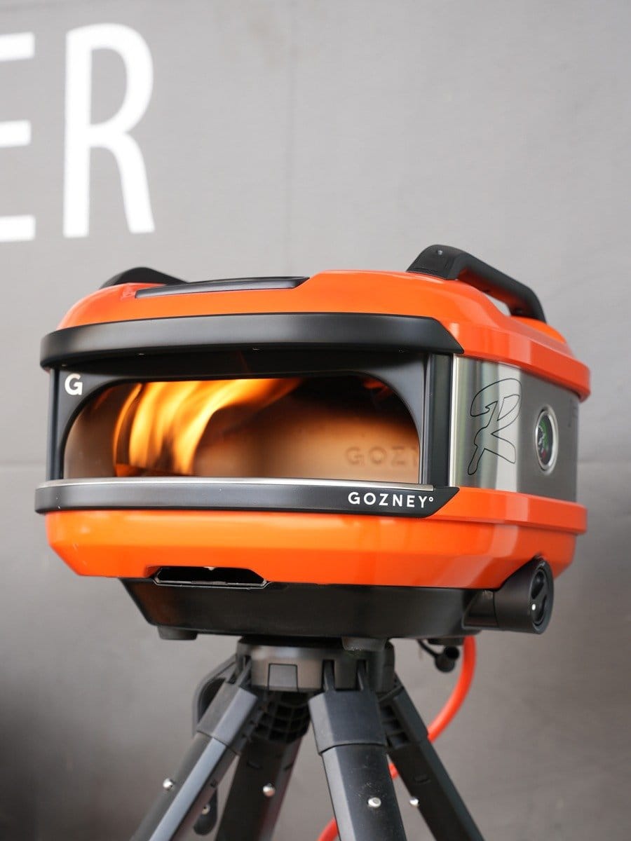 Ein tragbarer Gasbrenner auf einem Stativ, der eine Flamme erzeugt, mit orangefarbenem Gehäuse und schwarzem Griff, auf einer grauen Wand im Hintergrund.