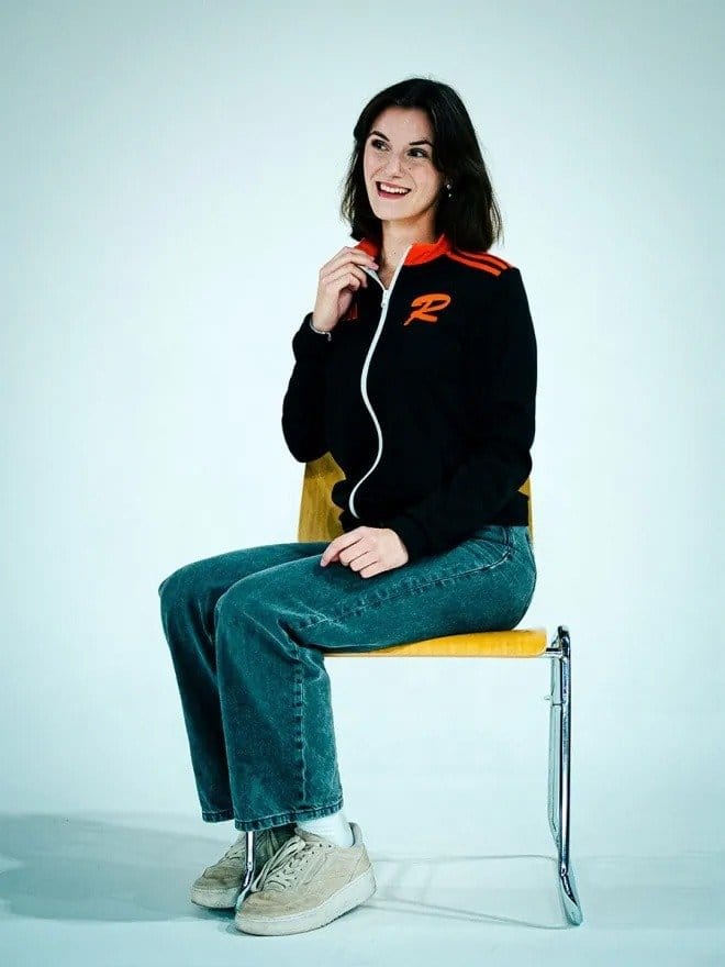 Junge Frau sitzt auf einem Stuhl, trägt eine schwarze Jacke mit roten Akzenten, blaue Jeans und weiße Sneakers, lächelt und schaut nach rechts.