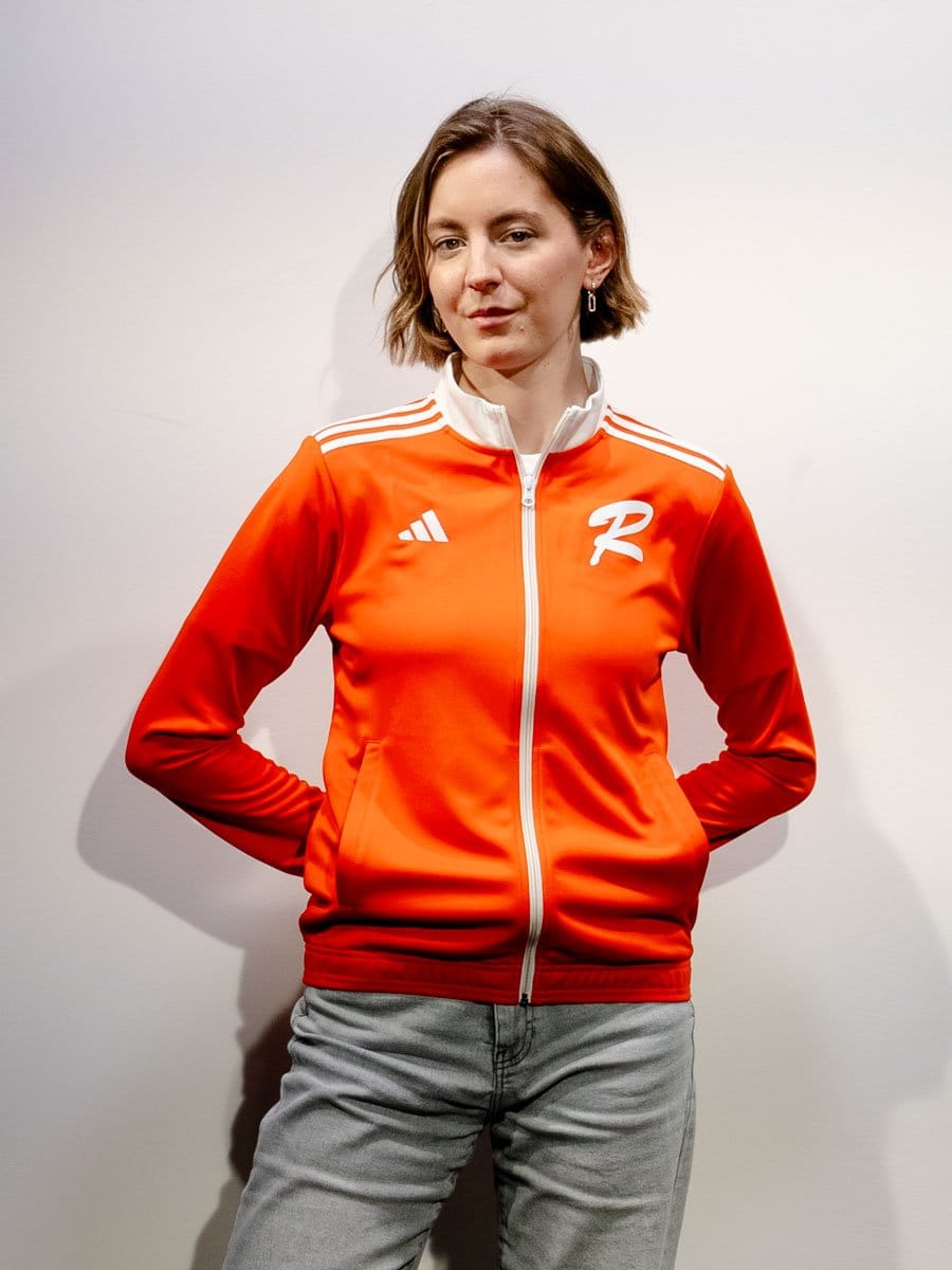 Junge Frau mit kurzen braunen Haaren trägt eine orange Sportjacke mit weißen Streifen und Logos, steht vor weißer Wand, Hände hinter dem Rücken, lächelt leicht.