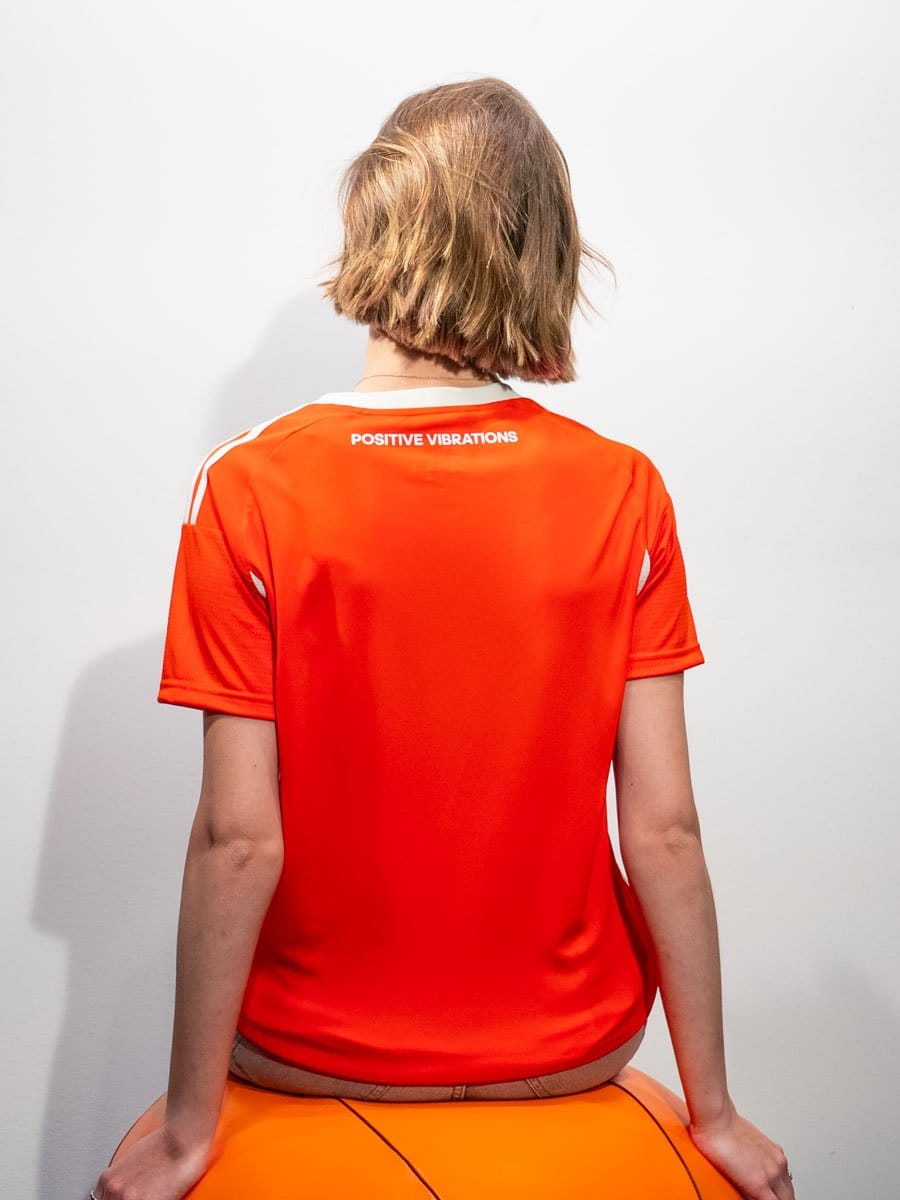 Rückenansicht einer Person mit kurzen, hellbraunen Haaren, die ein rotes Sportshirt mit weißen Streifen und dem Text "POSITIVE VIBRATIONS" trägt, auf einem orangefarbenen Ball sitzend.