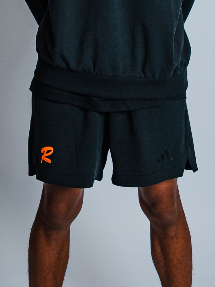 Schwarze Sportshorts mit orangeem und schwarzem Logo, getragen von einer Person mit dunkler Haut, gegen einen grauen Hintergrund.