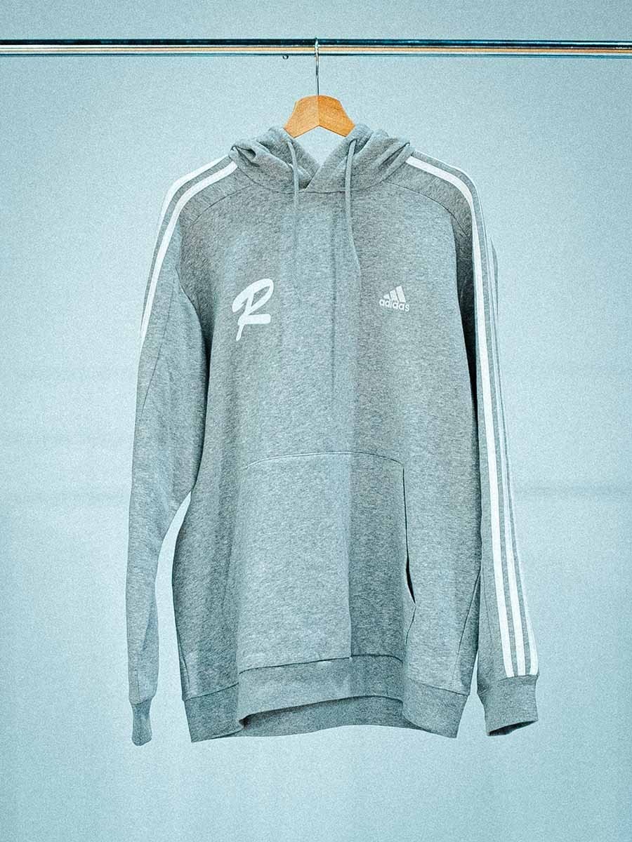 Grauer Adidas-Hoodie mit weißen Streifen an den Ärmeln, Logo auf Brust und Rücken, hängt an einem Holzkleiderbügel an einer Stange.