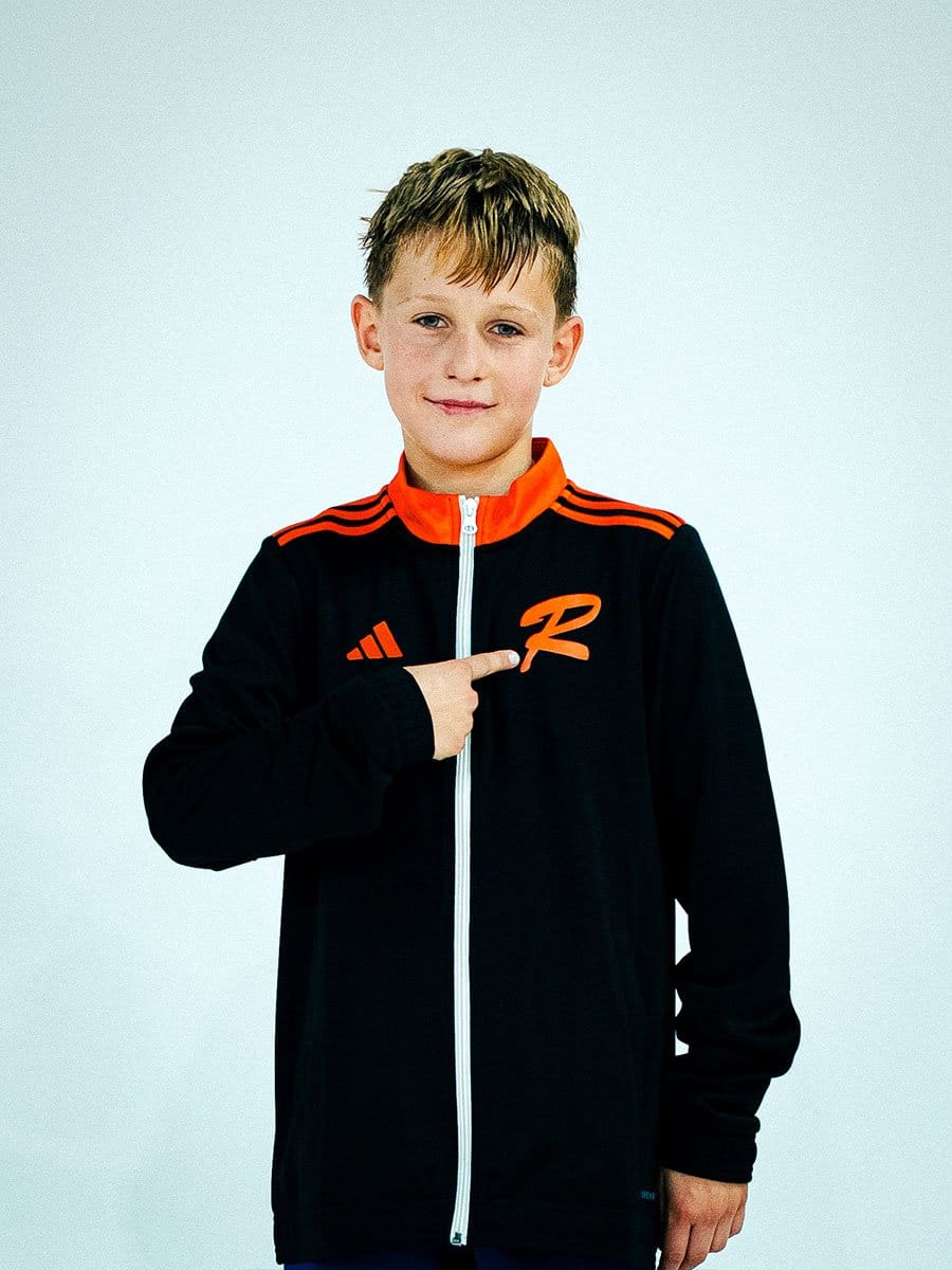 Junge trägt schwarze Sportjacke mit orangeem Reißverschluss, orange-gestreiften Schultern und einem orangefarbenen "R" auf der linken Brustseite, zeigt auf das Logo.