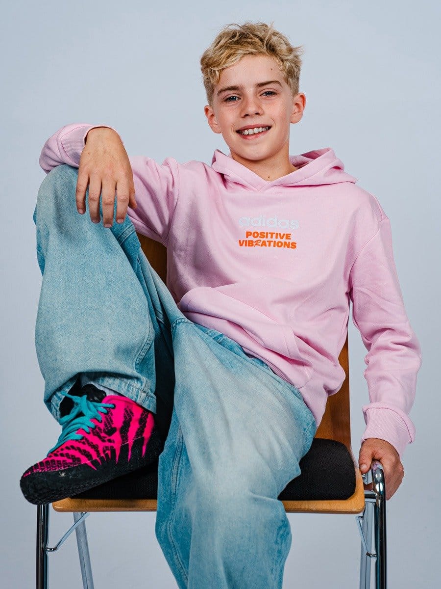 Junge Person sitzt auf einem Stuhl, trägt eine pinke Adidas-Hoodie mit dem Schriftzug "Positive Vibrations", blaue Jeans und bunte Sportschuhe, lächelt freundlich.