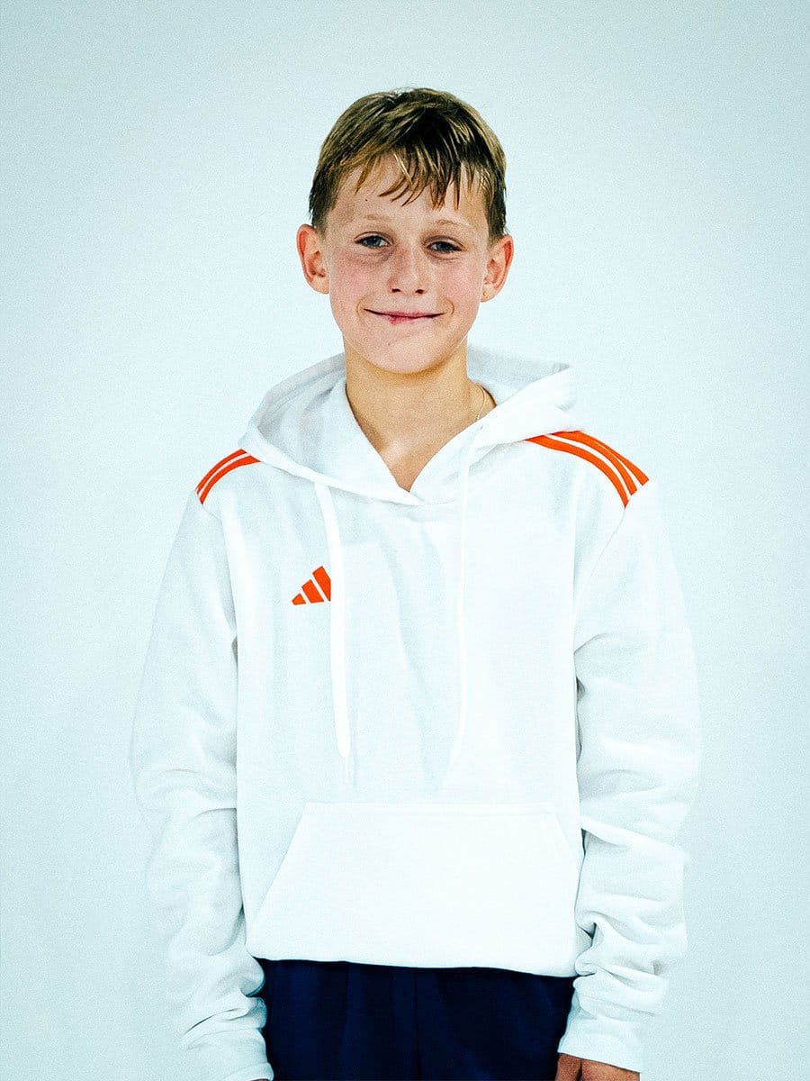 Junge Person trägt weiße Kapuzenjacke mit roten Streifen auf den Schultern und Adidas-Logo, steht vor hellem Hintergrund, lächelt leicht.