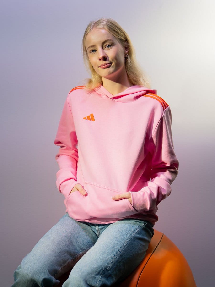 Junge Frau mit blondem Haar, trägt pinken Hoodie mit orange Streifen auf den Schultern, sitzt auf einem orangefarbenen Ball, vor hellem Hintergrund.