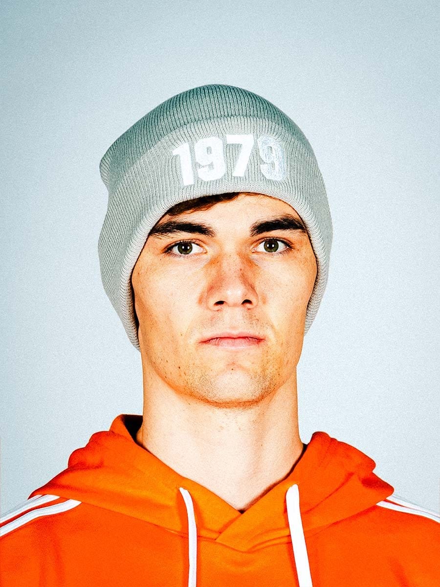 Junger Mann trägt grauen Beanie mit "1973" und orange Hoodie, blickt ernst in die Kamera, neutraler Hintergrund.