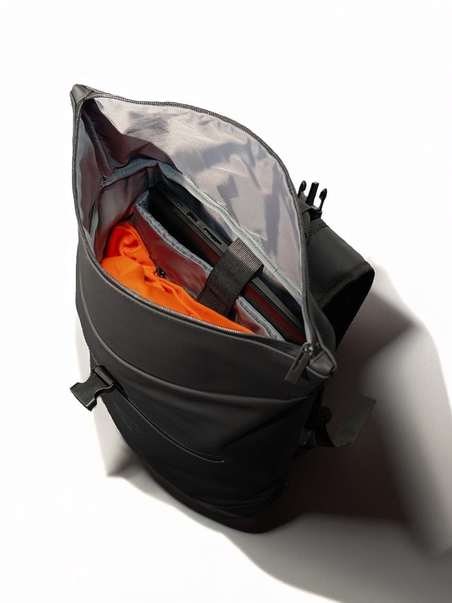 Schwarze Rucksack mit offenem Hauptfach, darin eine orange Jacke, ein Laptop und ein schwarzer Gegenstand. Der Rucksack liegt auf weißem Hintergrund.