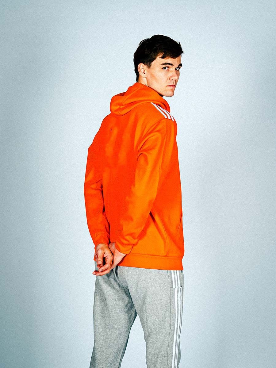 Junger Mann in orange Kapuzenjacke und grauen Jogginghose steht seitlich, blickt über die Schulter, Hände hinter dem Rücken, vor hellem Hintergrund.