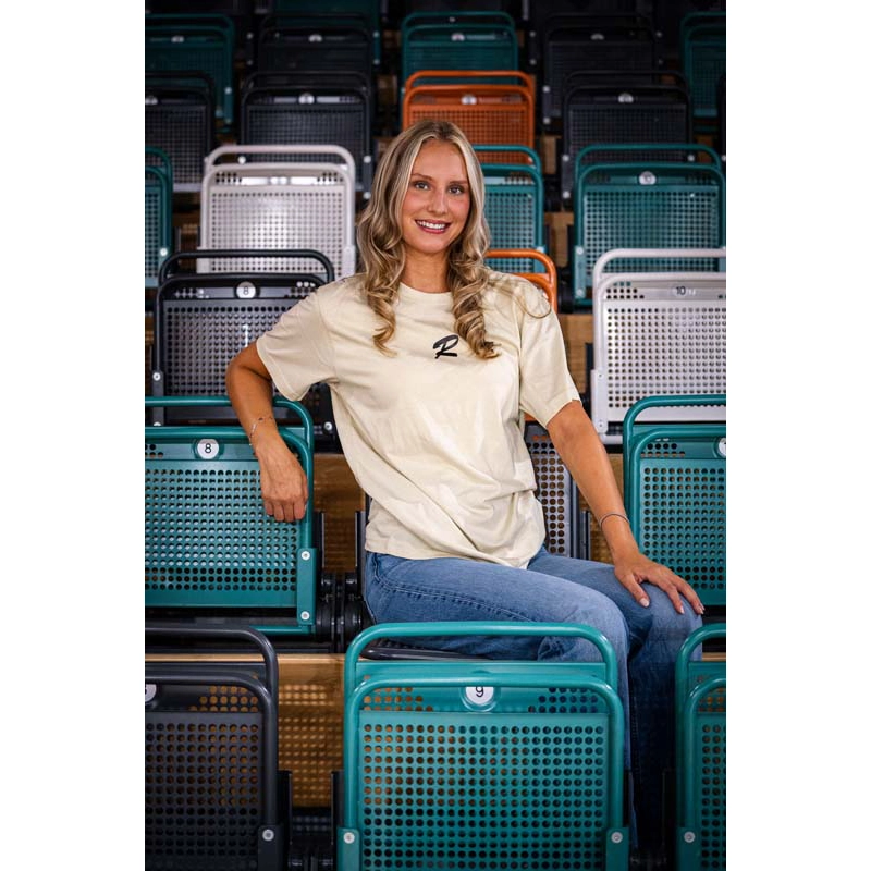 Frau sitzend mit Sandfarbenem T-Shirt und Rasta Logo auf der Brust in schwarz. 