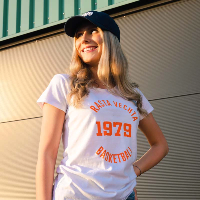 Frau mit Kappe im Damen T-Shirt "1979" in weiß mit Rasta Vechta Basketball  Schriftzug Kreisförmig und in der Mitte die Zahl 1979 als Aufdruck in orange.